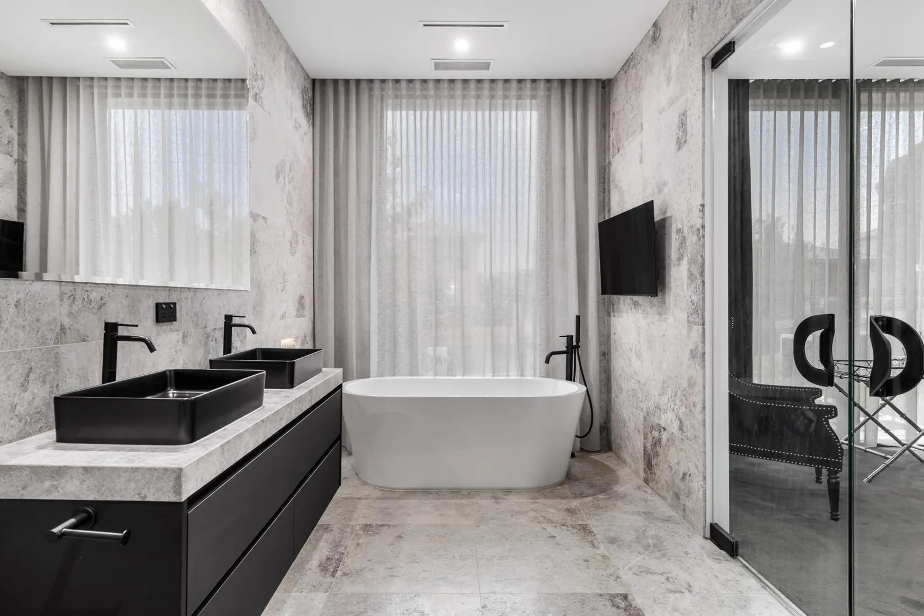 Freestanding bath between travertine walls, with a black double vanity and twin black basins