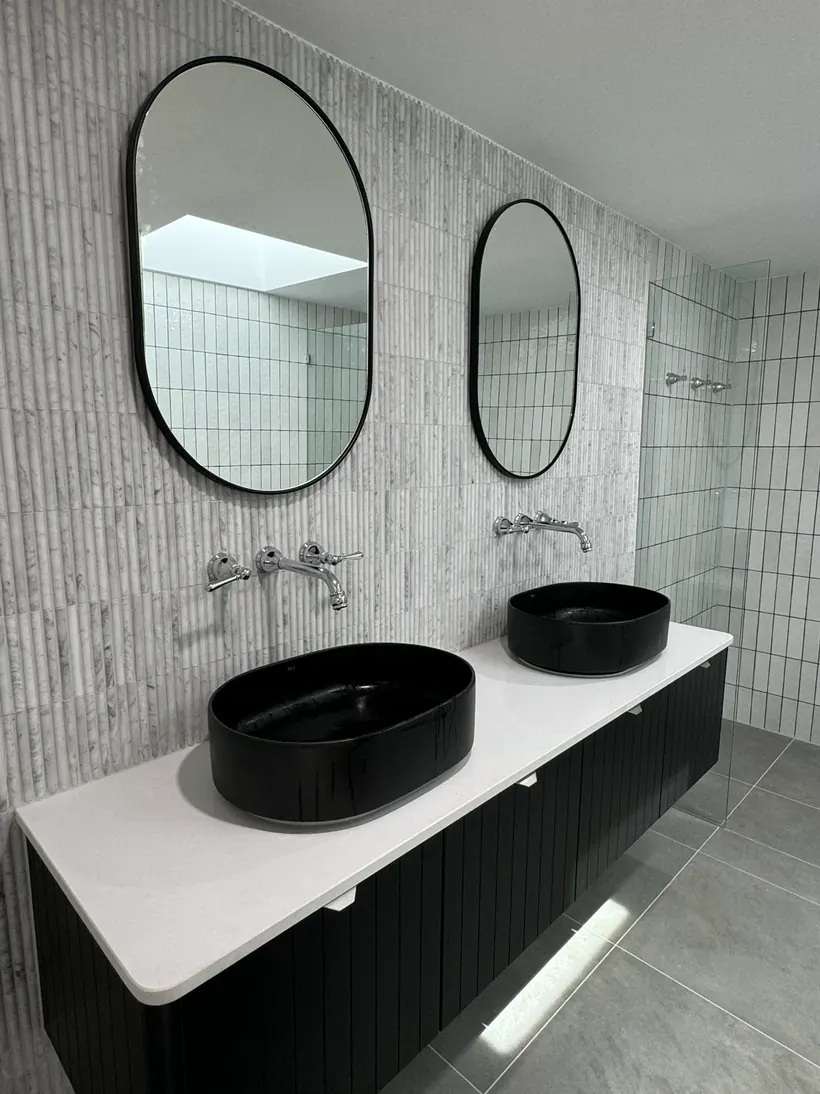 Twin oval mirrors above a black vanity with two black basins and white stacked tile